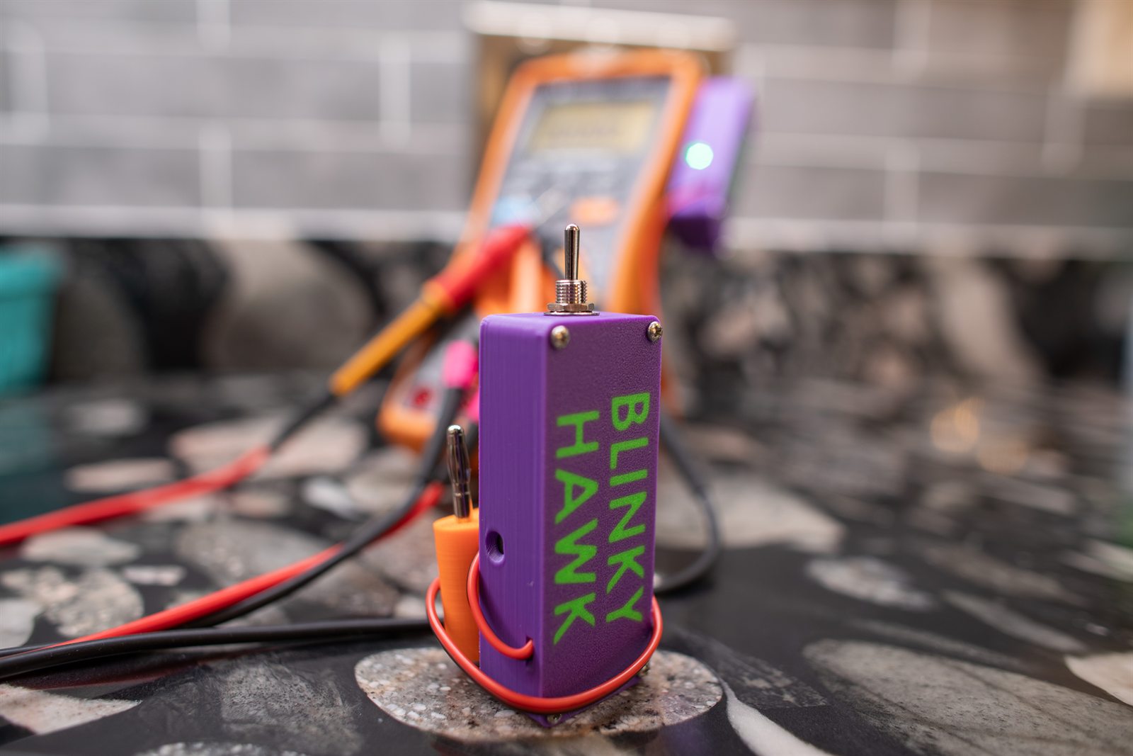 A purple Blinky Hawk standing upright on a stone countertop, the BLINKY HAWK name moulded into its side in green, with the metal toggle switch on top and a red lead looping around its base. An orange multimeter with a second purple unit attached is blurred behind it.