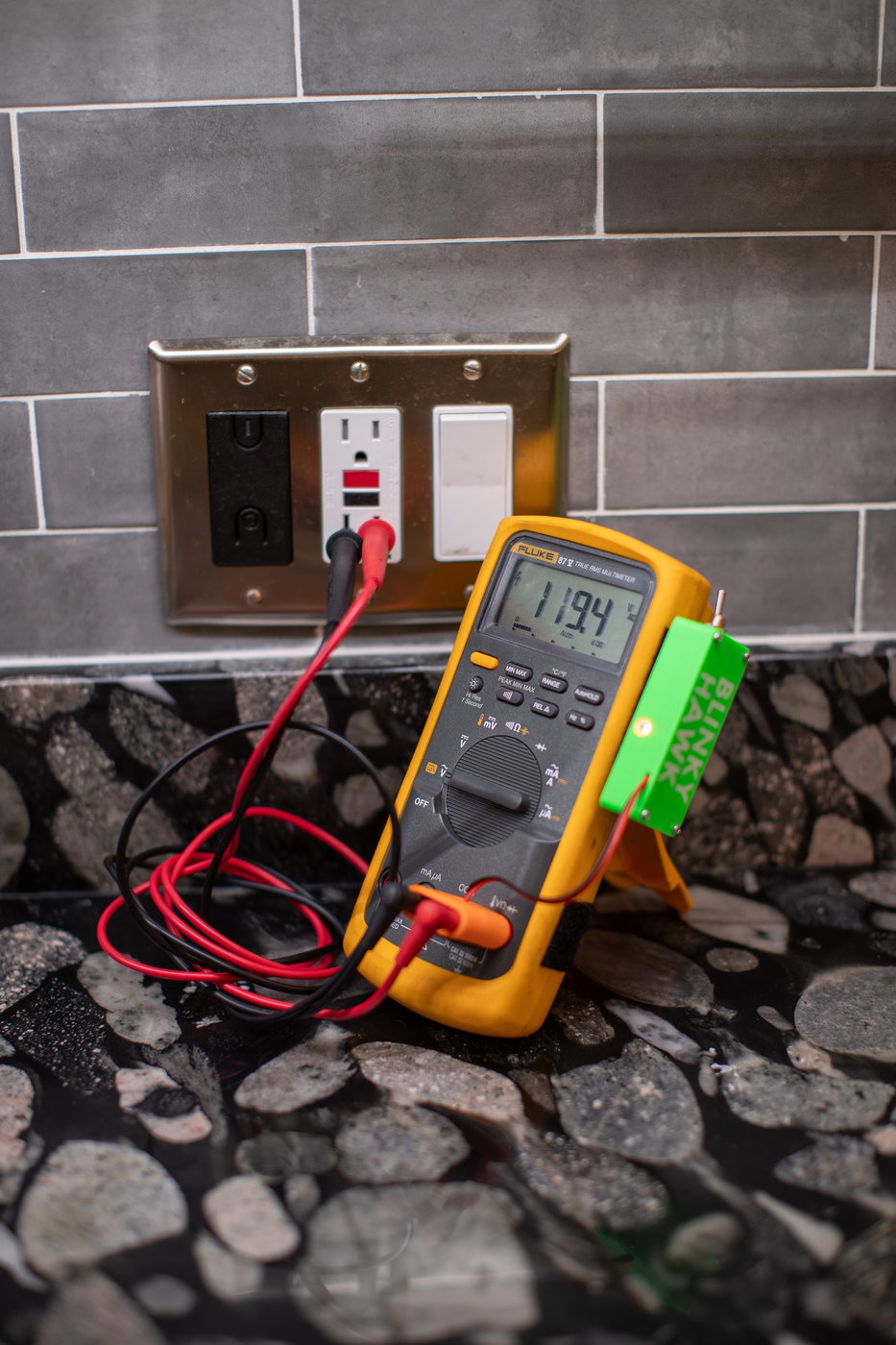 A Fluke 87V propped on a countertop with its probes in a wall outlet, reading 119.4 V AC. The Blinky Hawk clipped to its side is lit orange.