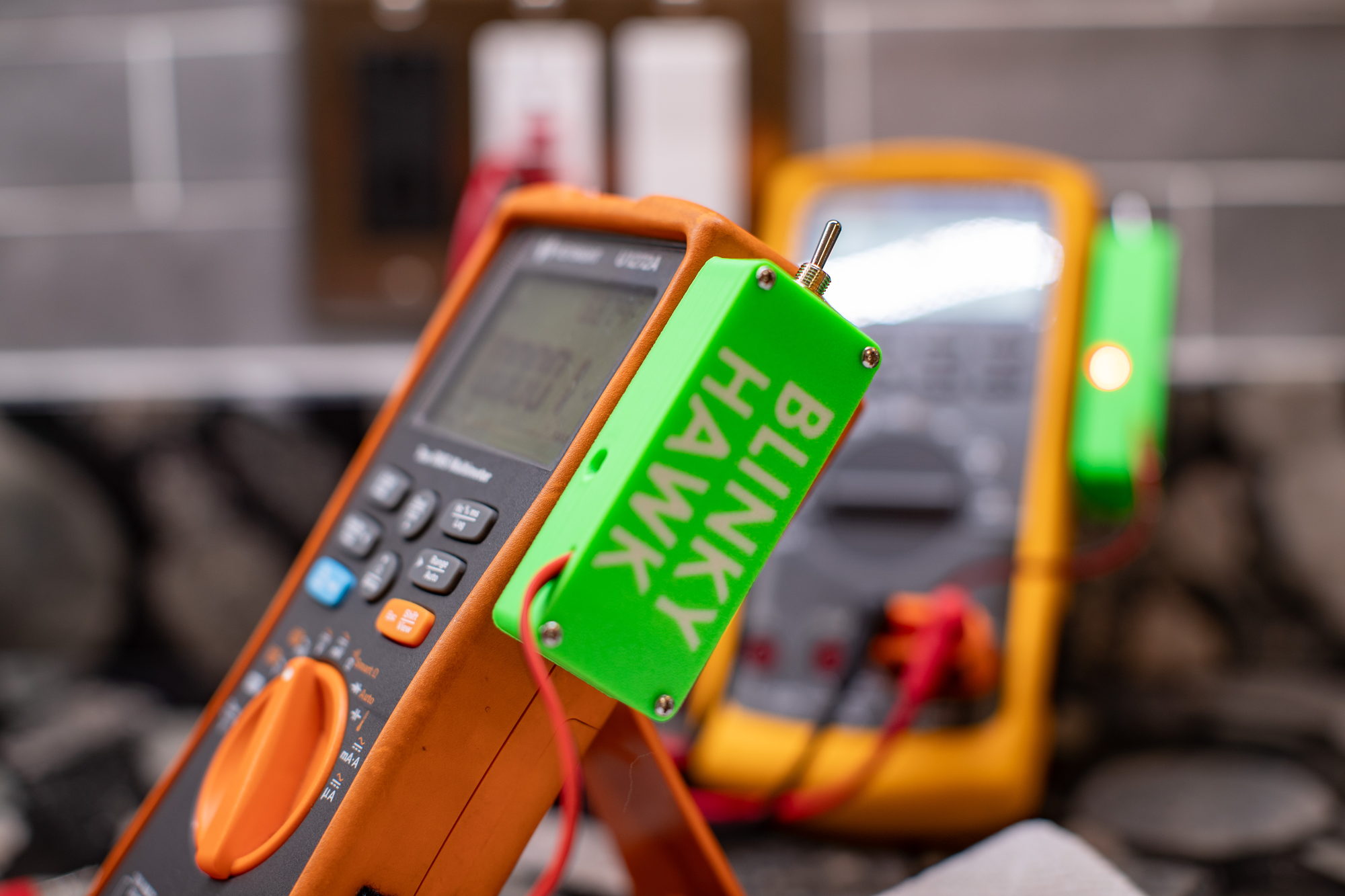 Close view of a green Blinky Hawk clipped to the side of an orange Keysight multimeter, the BLINKY HAWK name moulded into its housing. A second meter sits blurred behind it with its own detector glowing orange.